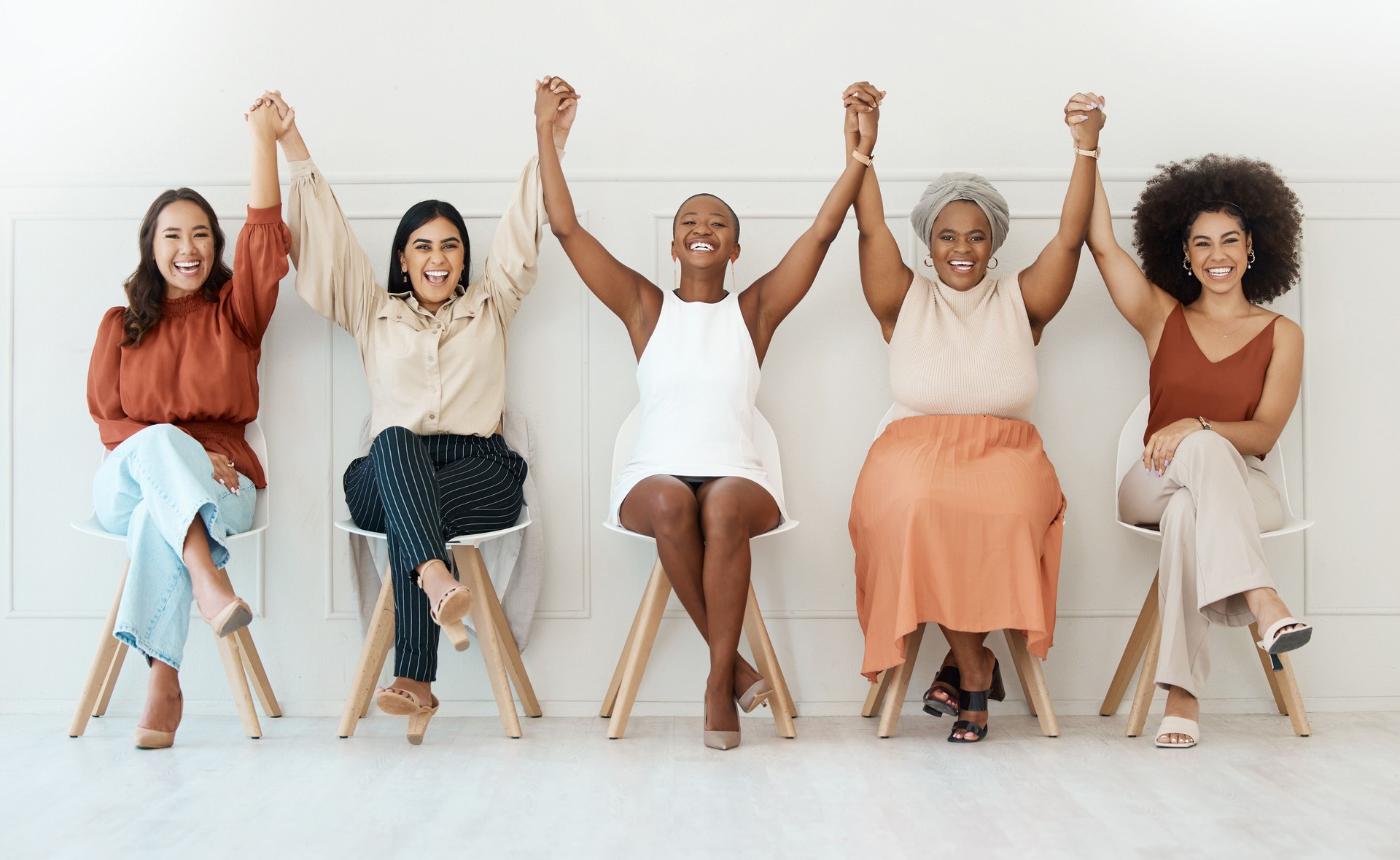 Group of five happy young businesswomen sitting against a wall in an office and holding hands in unity. Businesspeople smiling holding hands and sitting in a row together inside at work